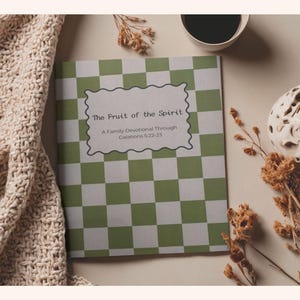 Puede incluir: Un libro devocional titulado "The Fruit of the Spirit" con un patrón de cuadros verdes y blancos. El libro está abierto, mostrando texto dentro de un marco decorativo. También hay una taza de café, un jarrón decorativo y flores secas.