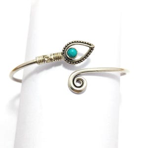 May include: An open-ended silver-toned metal cuff bracelet featuring a teardrop-shaped setting with a small, round, bright turquoise stone. The band includes a coiled wire detail near the teardrop and a spiral design at the opposite end.