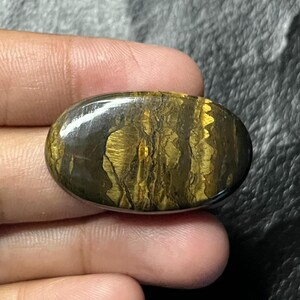 May include: An oval-shaped tiger's eye gemstone featuring a polished, smooth surface with characteristic chatoyant bands of golden-brown and dark brown colors that create a shimmering, layered appearance across the stone's face.