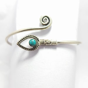 May include: A silver-toned metal cuff bracelet featuring a spiral end and a teardrop-shaped focal point. The teardrop element is decorated with a beaded border and contains a small, round, bright turquoise stone set in the center of the design.