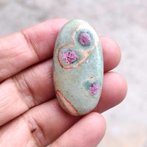 May include: An oval-shaped cabochon gemstone featuring a smooth, pale sage green surface with three distinct, embedded deep pink ruby inclusions, each surrounded by a subtle, light brown circular halo pattern within the stone's natural texture.
