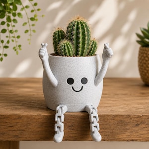 May include: A light grey speckled planter shaped like a character with a smiling face, two arms raised in a heart-hand gesture, and two dangling articulated legs. The planter holds a green, multi-stemmed cactus with visible vertical spines.