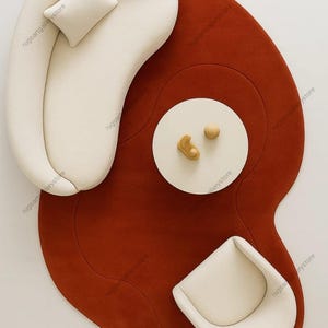 May include: An overhead view of a modern living room setup. A large, rust-colored, organically shaped rug anchors the space. A white, curved sofa with a matching pillow, a small round table with decorative objects, and a white chair complete the arrangement.