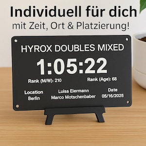 Könnte beinhalten: Eine schwarze rechteckige Plakette mit weißem Text, die "HYROX DOUBLES MIXED" und eine Zeit von 1:05:22 anzeigt. Sie enthält Rang, Ort, Namen und ein Datum. Die Plakette wird von einem schwarzen Ständer getragen.