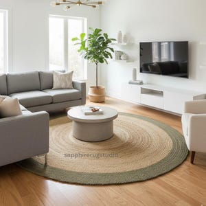 May include: A round jute rug with a natural tan center and a sage green border. The rug is in a living room setting with gray furniture, a coffee table, and a potted plant. The rug is a decorative floor covering.