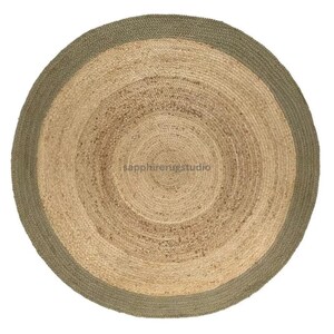 May include: A round jute rug with a natural tan center and a sage green border. The rug has a woven texture and a circular design, suitable for a living room or bedroom. The rug's diameter is approximately 60 inches.