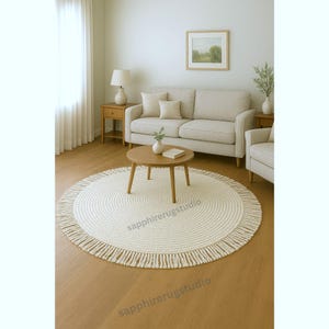 May include: A circular, cream-coloured rug with a fringed border is placed on a wooden floor. The rug is in a living room, with a wooden coffee table and a beige sofa in the background. The room is bright with natural light.