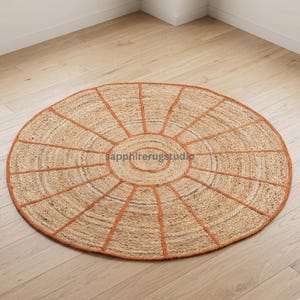 May include: A round jute rug with a sunburst design. The rug features a natural tan color with a burnt orange border and radiating lines. The rug is placed on a light wood floor.