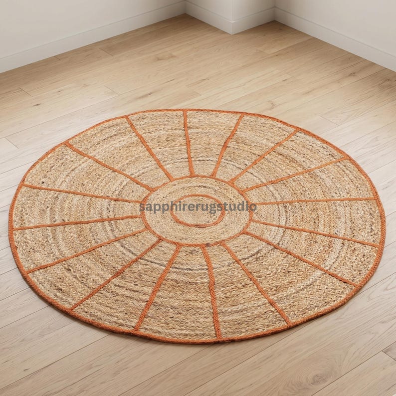 May include: A round jute rug with a sunburst design. The rug features a natural tan color with a burnt orange border and radiating lines. The rug is placed on a light wood floor.