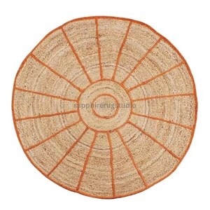 May include: Round jute rug with a sunburst design. The rug features a natural beige color with orange stitching that outlines the circular shape and radiating lines. The rug has the text "sapphirerugstudio" in the center.