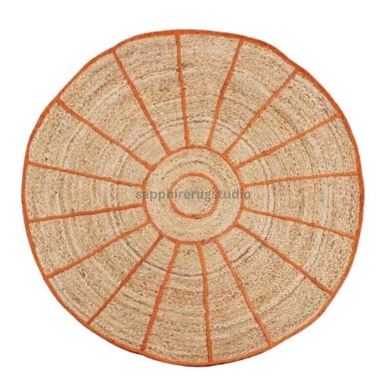 May include: Round jute rug with a sunburst design. The rug features a natural beige color with orange stitching that outlines the circular shape and radiating lines. The rug has the text "sapphirerugstudio" in the center.