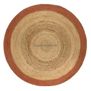 May include: A round jute rug with a natural beige center and a rust-colored border. The rug has a woven texture and a concentric circular design. The rug is suitable for indoor use.