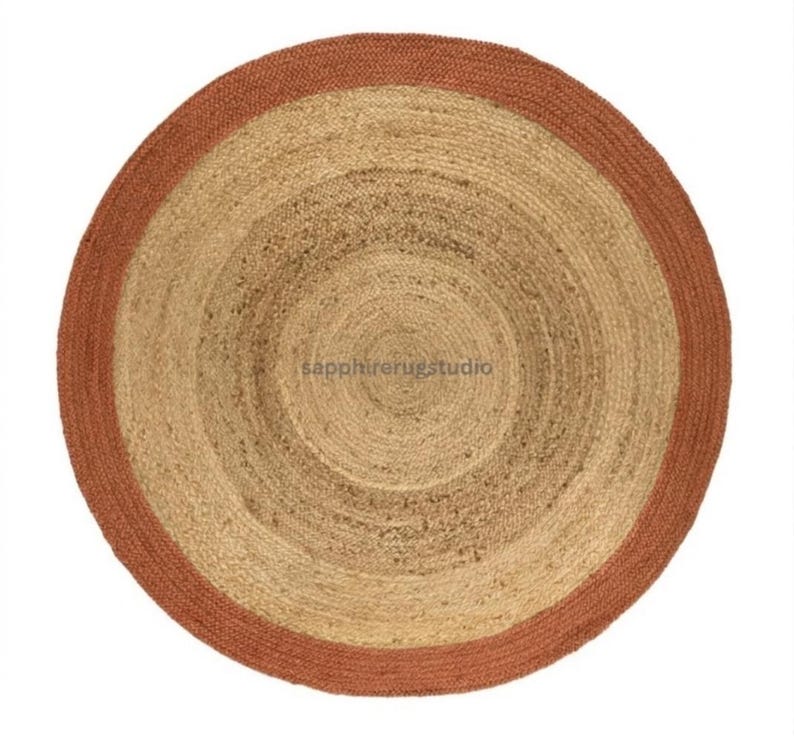 May include: A round jute rug with a natural beige center and a rust-colored border. The rug has a woven texture and a concentric circular design. The rug is suitable for indoor use.