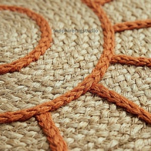 May include: Close-up of a woven jute rug with a geometric design. The rug features a natural beige color with a burnt orange rope-like trim. The text "sapphirerugstudio" is visible in the upper left corner.
