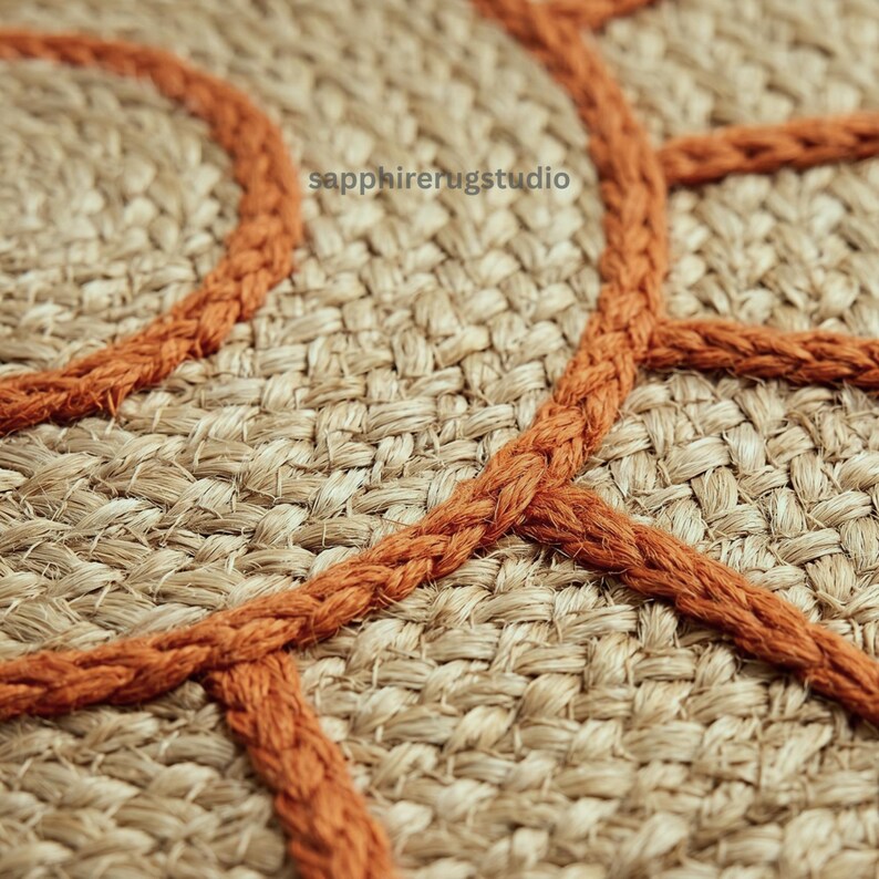 May include: Close-up of a woven jute rug with a geometric design. The rug features a natural beige color with a burnt orange rope-like trim. The text "sapphirerugstudio" is visible in the upper left corner.