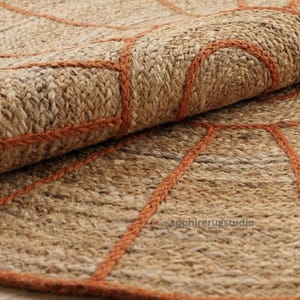 May include: Close-up of a rolled-up jute rug with a geometric design. The rug is a natural tan color with rust-colored stitching outlining the pattern. The texture of the rug is visible, showing the woven fibers.