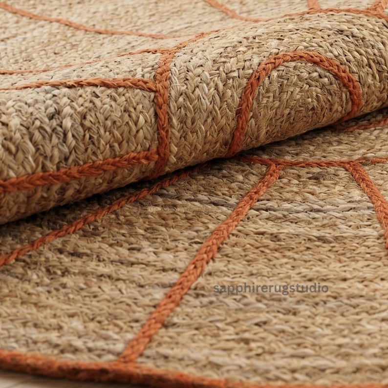 May include: Close-up of a rolled-up jute rug with a geometric design. The rug is a natural tan color with rust-colored stitching outlining the pattern. The texture of the rug is visible, showing the woven fibers.