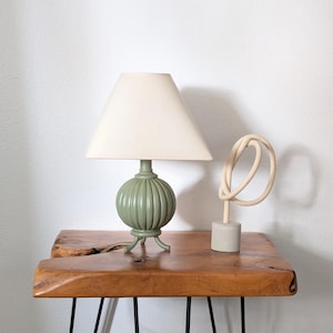 Peut inclure: Une lampe de table avec une base sphérique côtelée vert sauge et un abat-jour conique crème, placée à côté d'une sculpture abstraite beige présentant une forme en boucle continue sur une base cylindrique, posée sur une surface en bois.