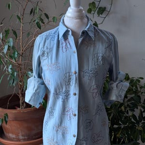 May include: Pale blue button-down shirt featuring embroidered leaf patterns in multiple colours. The long-sleeved shirt is displayed on a mannequin. The sleeves are rolled up. The shirt has a collar and is buttoned up.
