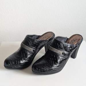 May include: A pair of black leather clogs with a woven texture and high heels. Each shoe has a silver-toned strap with a decorative design. The brand name "NEW TRA" is visible on the inner sole.