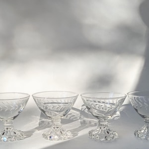 Op de afbeelding: Vier heldere champagnecoupeglazen met een bladerenpatroon. Elk glas heeft een ronde kom en staat op een decoratieve voet. De glazen zijn in een rij gerangschikt tegen een witte achtergrond.