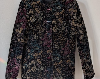 May include: A black floral print jacket with long sleeves and a button-up front. The jacket features a pattern of flowers in shades of pink, tan, and teal. The jacket is hanging on a white hanger.