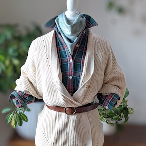 May include: Cream-colored, chunky knit cardigan layered over a plaid shirt with red and green accents. A brown leather belt cinches the waist. A patterned scarf and a small pin add detail to the outfit. The outfit is displayed on a mannequin.