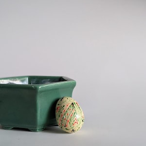 May include: A green ceramic planter with a square shape and a decorative egg. The egg is painted with a colorful floral design in red, green, and yellow. The planter is a solid, dark green color.