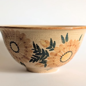 May include: A beige ceramic bowl featuring a hand-painted floral pattern in shades of light brown, yellow, and dark green. The design includes large stylized flowers with dotted centers and leafy stems encircling the exterior of the bowl.