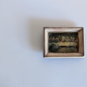 May include: A miniature rectangular wall art piece featuring a reproduction of The Last Supper painting. The artwork is encased in a light-colored, distressed wooden frame with a textured finish, highlighting the detailed religious scene within.