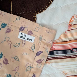 Puede incluir: Primer plano de una sección de edredón con tela color melocotón con un estampado de hojas y el texto "BOB WILLIAMS" en una etiqueta blanca. El edredón también incluye secciones marrones y blancas.