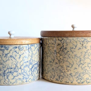 May include: Two cylindrical ceramic canisters with a cream-coloured base and a blue sponge-painted pattern. Each canister features a flat, circular wooden lid with a small, centred white knob handle. The canisters are of two different sizes.