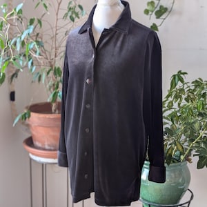 Puede incluir: Una camisa gris oscuro con botones, mangas largas y cuello, exhibida en un maniquí blanco. La camisa tiene una textura suave y aterciopelada y está abotonada. El fondo incluye plantas en macetas.