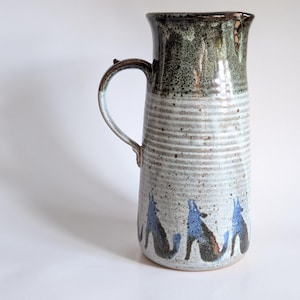 Può includere: Una brocca in ceramica con bordo smaltato verde scuro e corpo grigio chiaro con texture a coste orizzontali. La parte inferiore è decorata con un motivo ripetitivo di sagome di lupi ululanti blu e neri.