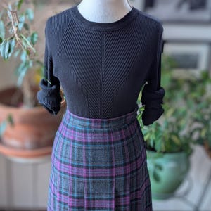 May include: A black jumper and a pleated plaid skirt. The skirt has a grey base with purple and teal plaid lines. The jumper has a ribbed texture and a crew neck. The outfit is displayed on a mannequin.