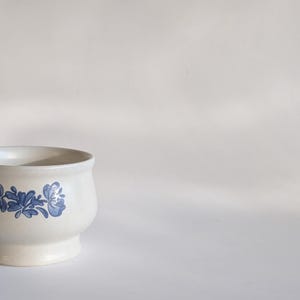 Puede incluir: Jardinera de cerámica color crema con un diseño floral azul. La jardinera tiene un cuerpo redondeado y una base de pedestal. El patrón floral azul se repite alrededor del cuerpo de la jardinera. Adecuada para uso interior o exterior.