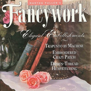 May include: The cover of a vintage "Fancywork" magazine features the title in large white letters. The cover includes text about elegant embellishments, embroidery, and a free pattern. Pink roses and antique books are also visible.