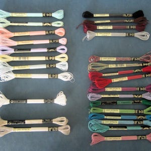 May include: Assortment of embroidery floss in various colors, including pastels, reds, and dark hues. Each skein is labeled with "Light Effects" and a product code. The floss is neatly arranged on a blue surface, ideal for craft projects.