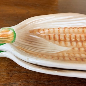 May include: A ceramic corn-shaped serving dish. The dish is painted with realistic details, featuring a tan husk, brown kernels, and green accents. The dish is designed to resemble an ear of corn.