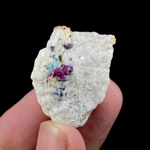 Op de afbeelding: Een mineraalmonster met een witte matrix, met clusters van rode, blauwe en groene kristallen. De ruwe textuur van de steen is zichtbaar. De achtergrond is zwart.