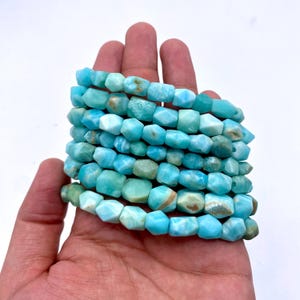 May include: A stack of light blue larimar gemstone beads. The beads have a faceted, irregular shape and a mix of light blue and white colors, with some brown inclusions. The beads are held in a hand against a white background.