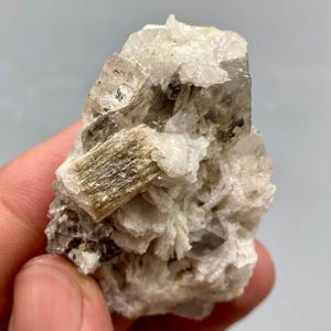 May include: A natural mineral specimen with a cluster of clear, crystalline formations and a central, elongated, tan-coloured crystal. The mineral has a rough, textured surface with a mix of white and grey hues.