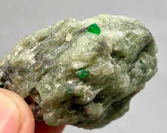 May include: A rough, irregularly shaped, light green rock with several small, vibrant green emerald crystals embedded within. The rock has a textured surface and appears to be a raw, natural mineral specimen.