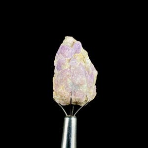 May include: A rough, natural gemstone with a light purple hue, held by a metal tool against a black background. The stone has a textured surface and a mix of light purple and beige colors, showcasing its raw, unpolished state.