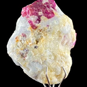 May include: A raw mineral specimen featuring a prominent, deep magenta-colored crystal embedded within a textured, off-white and cream-colored matrix. The stone displays irregular, jagged edges and a crystalline surface structure with small, dark inclusions.