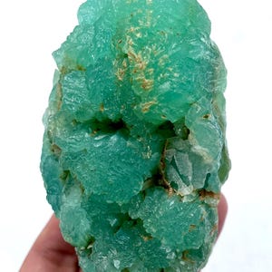 May include: A vibrant, raw, teal-colored mineral specimen. The crystalline structure has a rough, textured surface with areas of light brown. The mineral is held against a white background, highlighting its natural beauty.