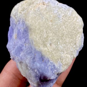 May include: A rough, natural mineral specimen with a light lavender-blue hue on one side and a creamy white color on the other. The stone has a textured, uneven surface and is held in a hand against a dark background.