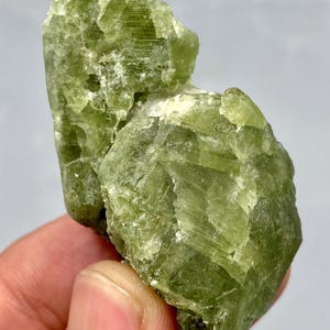 May include: A raw, natural peridot gemstone with a vibrant green hue. The crystal structure is visible, showcasing the mineral's rough, unpolished texture. The peridot is held against a neutral background, highlighting its color and form.