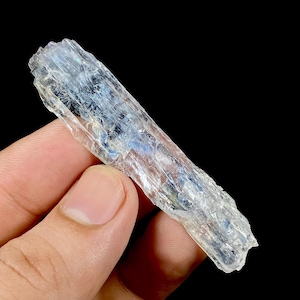 May include: A translucent, elongated crystal with a light blue hue and a rough, textured surface. The crystal is held against a black background, showcasing its natural form and color variations. The crystal appears to be a mineral specimen.