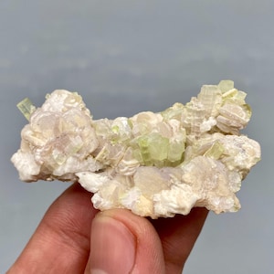 May include: A cluster of pale green and white crystals. The crystals have a layered appearance, with some sections showing a translucent quality. The overall structure is irregular and textured, with a mix of rounded and angular forms.